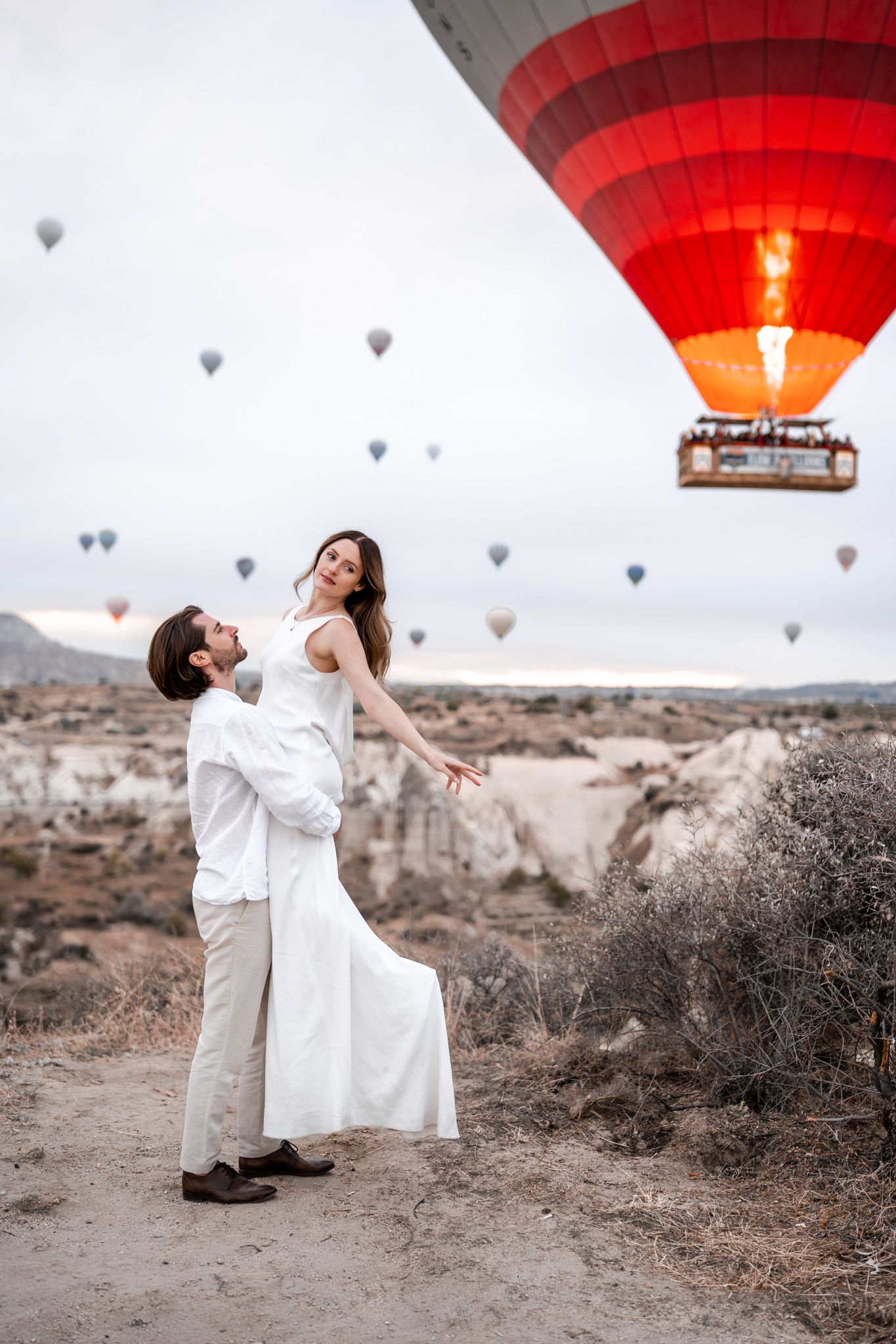 A romantic couple embraces beneath hot air balloons in a desert landscape.