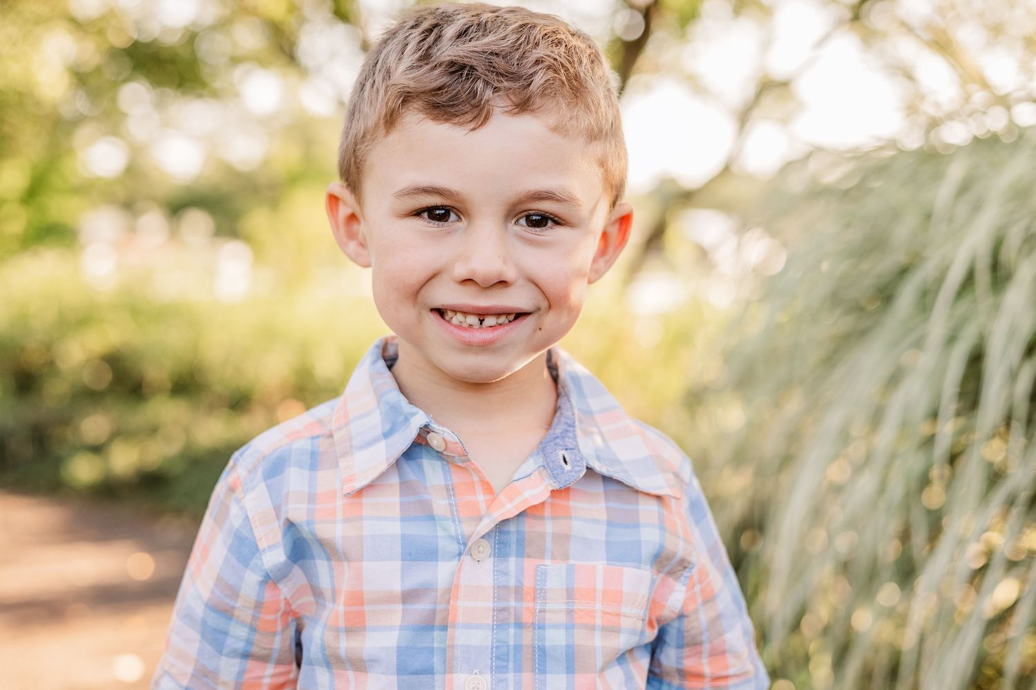 Child in plaid button-down shirt smiles warmly at camera outdoors.