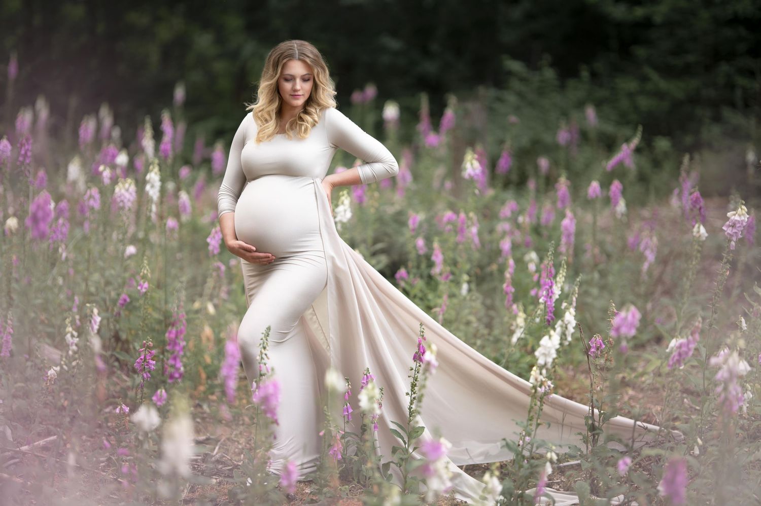 zwangere vrouw poseert tussen vingerhoedskruid in jurk met lange chiffon sleep
