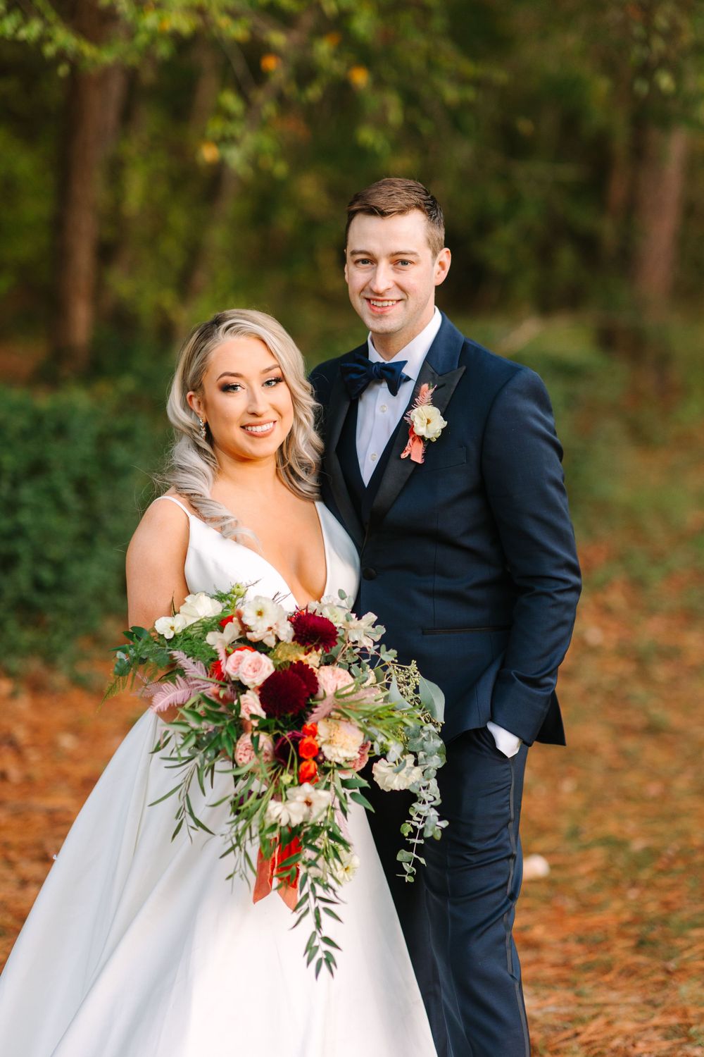 Couple poses with stunning burgundy and blush wedding bouquet in autumn setting.