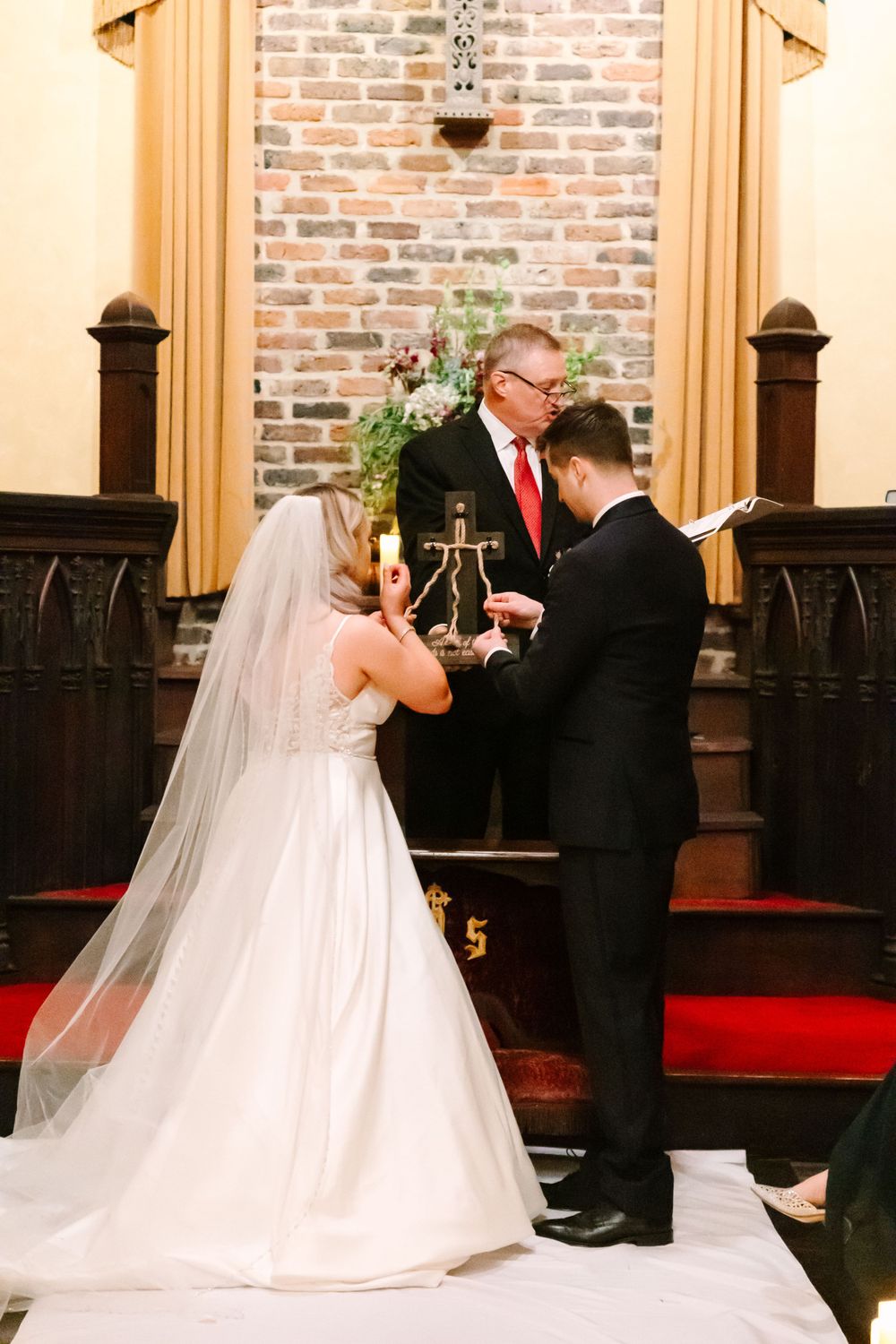 A wedding ceremony in a brick church with wooden columns and red carpet during the exchange of vows.