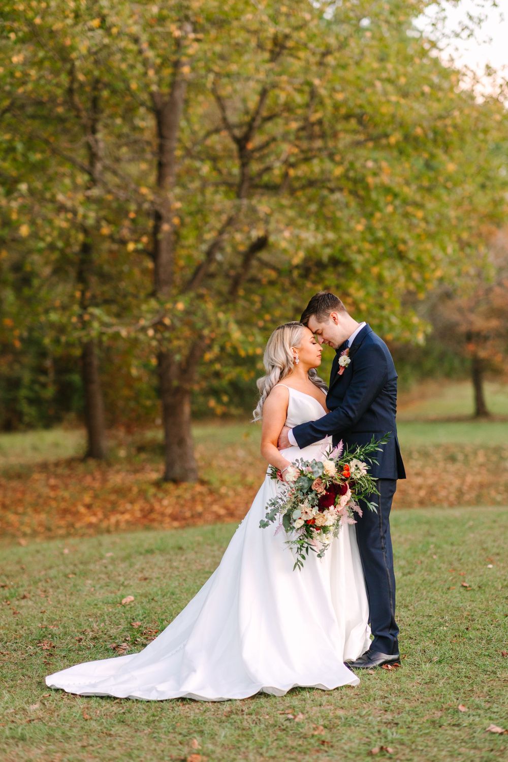 A romantic autumn wedding photo with a couple embracing on a lawn surrounded by fall foliage.