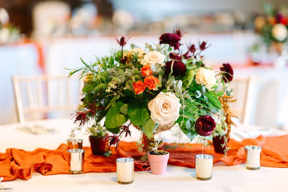Elegant table centerpiece with burgundy and orange flowers surrounded by votive candles.