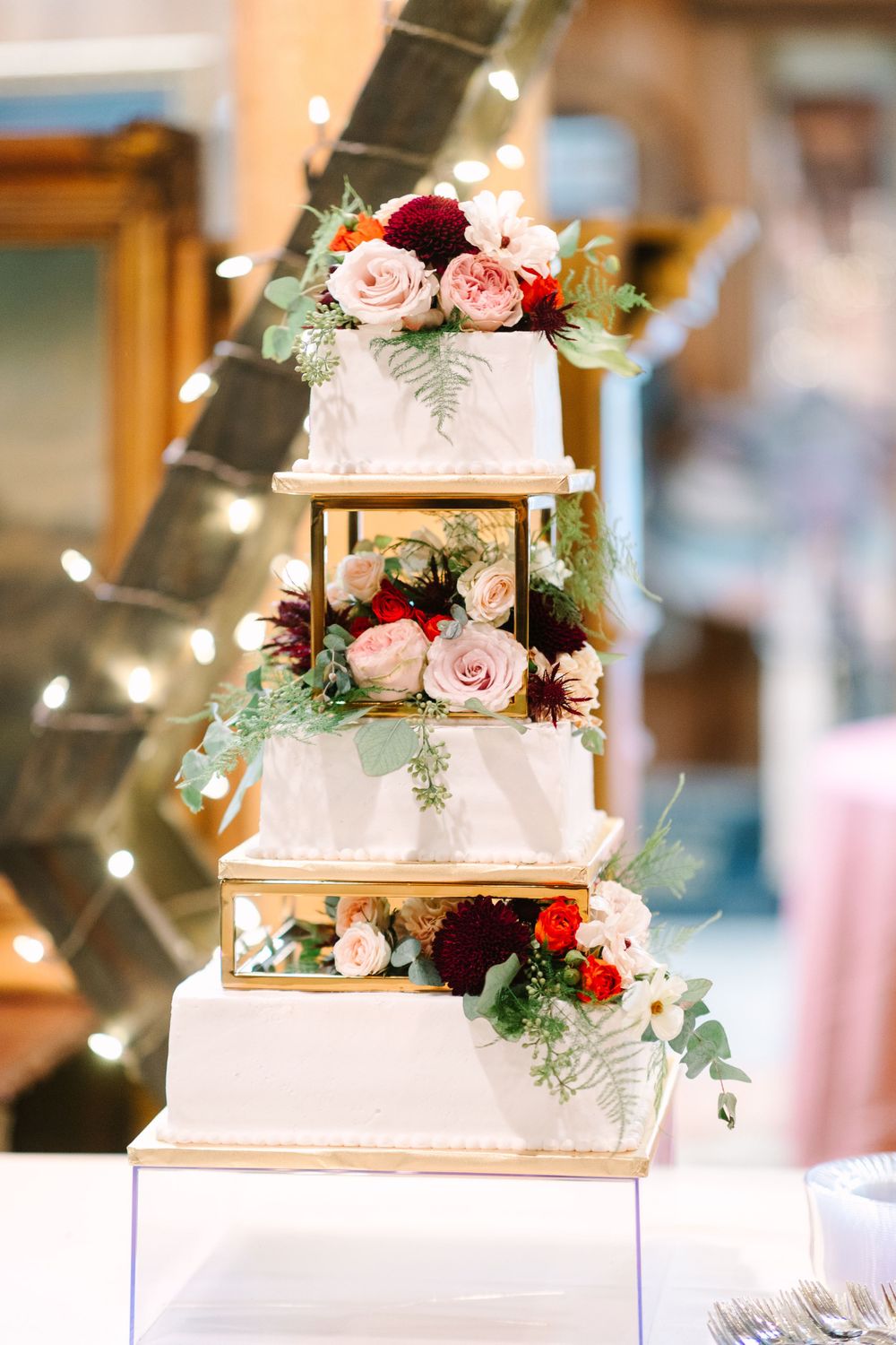 Three-tier white wedding cake decorated with pink and burgundy roses on gold stands.