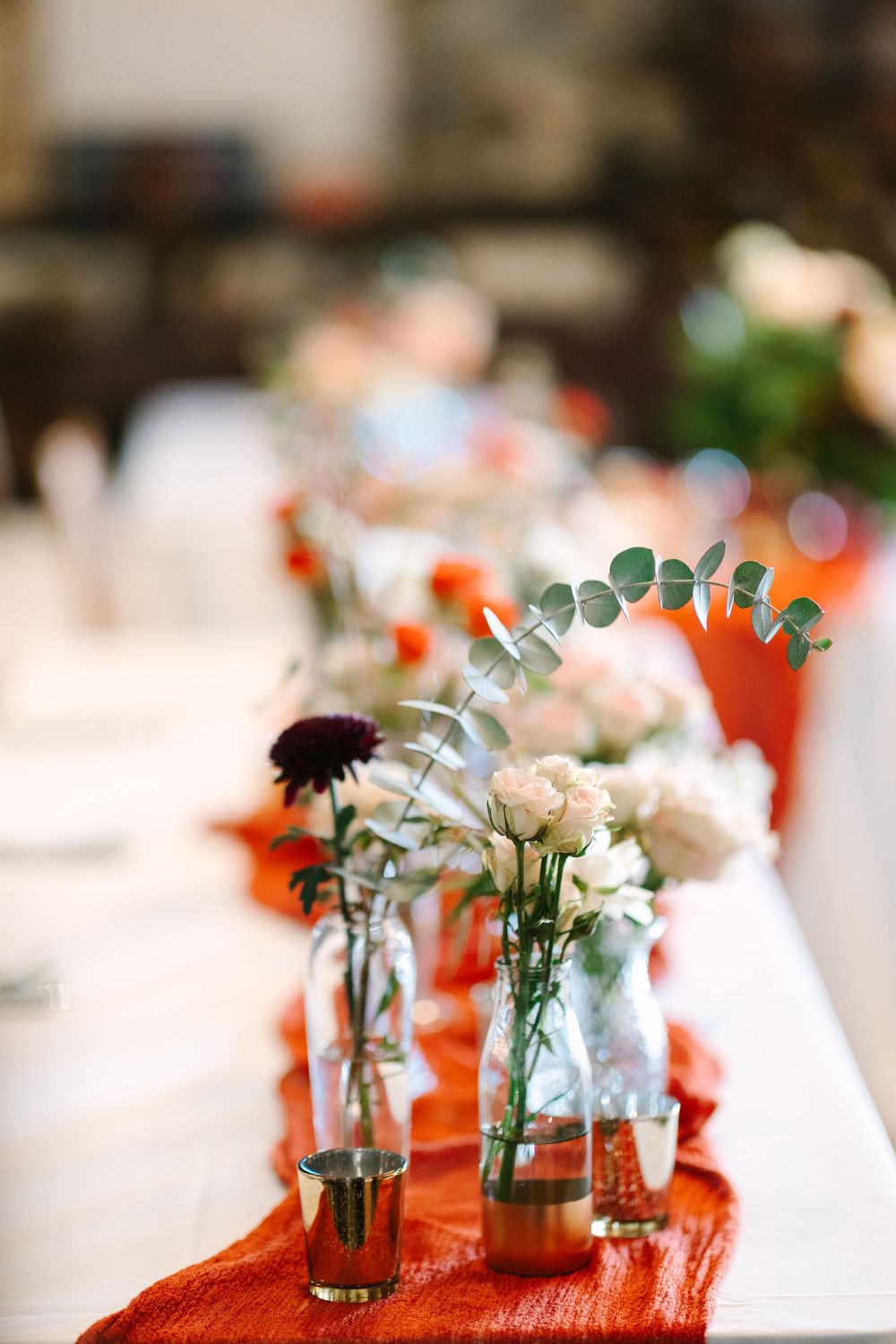 Small glass vases with delicate flowers and eucalyptus arranged on a white tablecloth with orange accents.