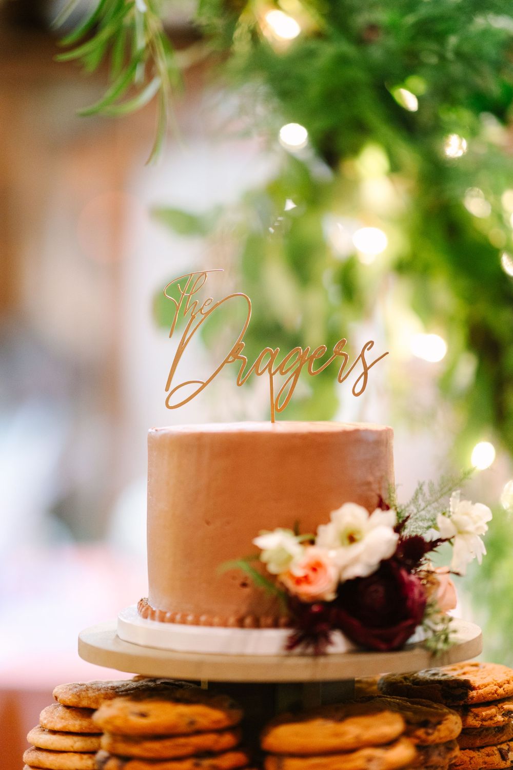 Small peach-colored cake with 'Rogers' topper decorated with fresh flowers and stacked on cookies.