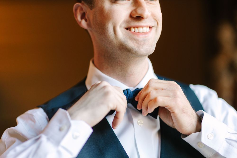 Person adjusting white tie while wearing navy vest and white dress shirt.