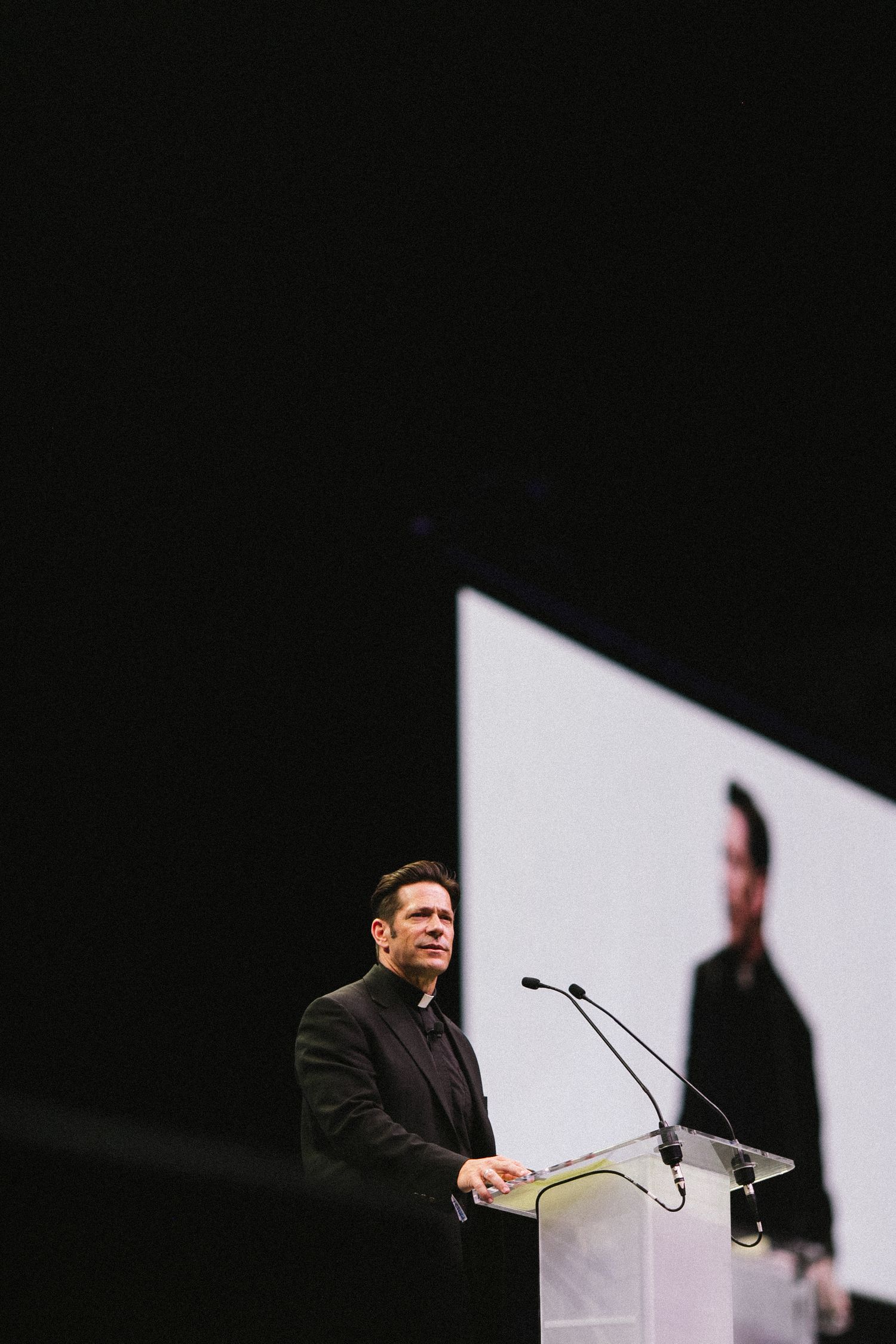 Speaker at podium against dark background with projection screen visible.