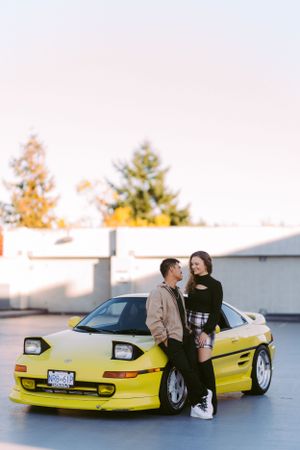 Yellow sports car parked outdoors with couple posing against it at sunset.