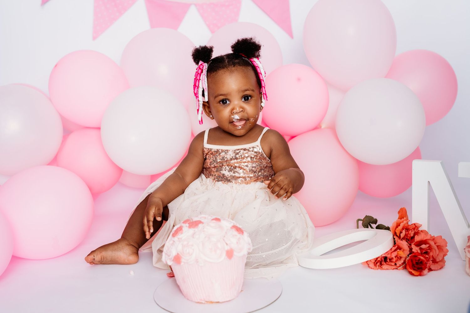 Baby in sparkly dress at cake smash photo session with pink balloon backdrop.