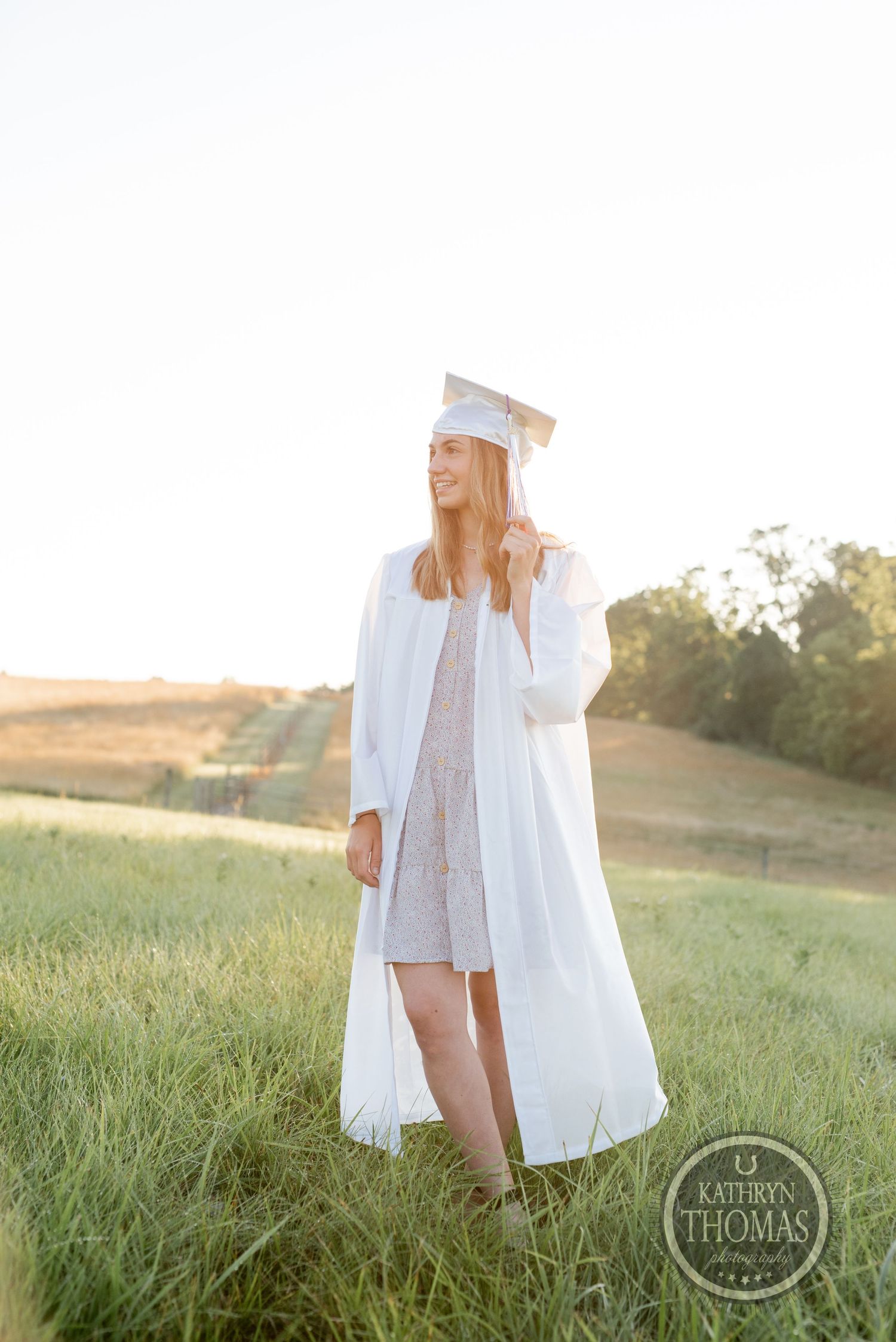 Graduate in white academic regalia poses in sun-drenched field during golden hour.