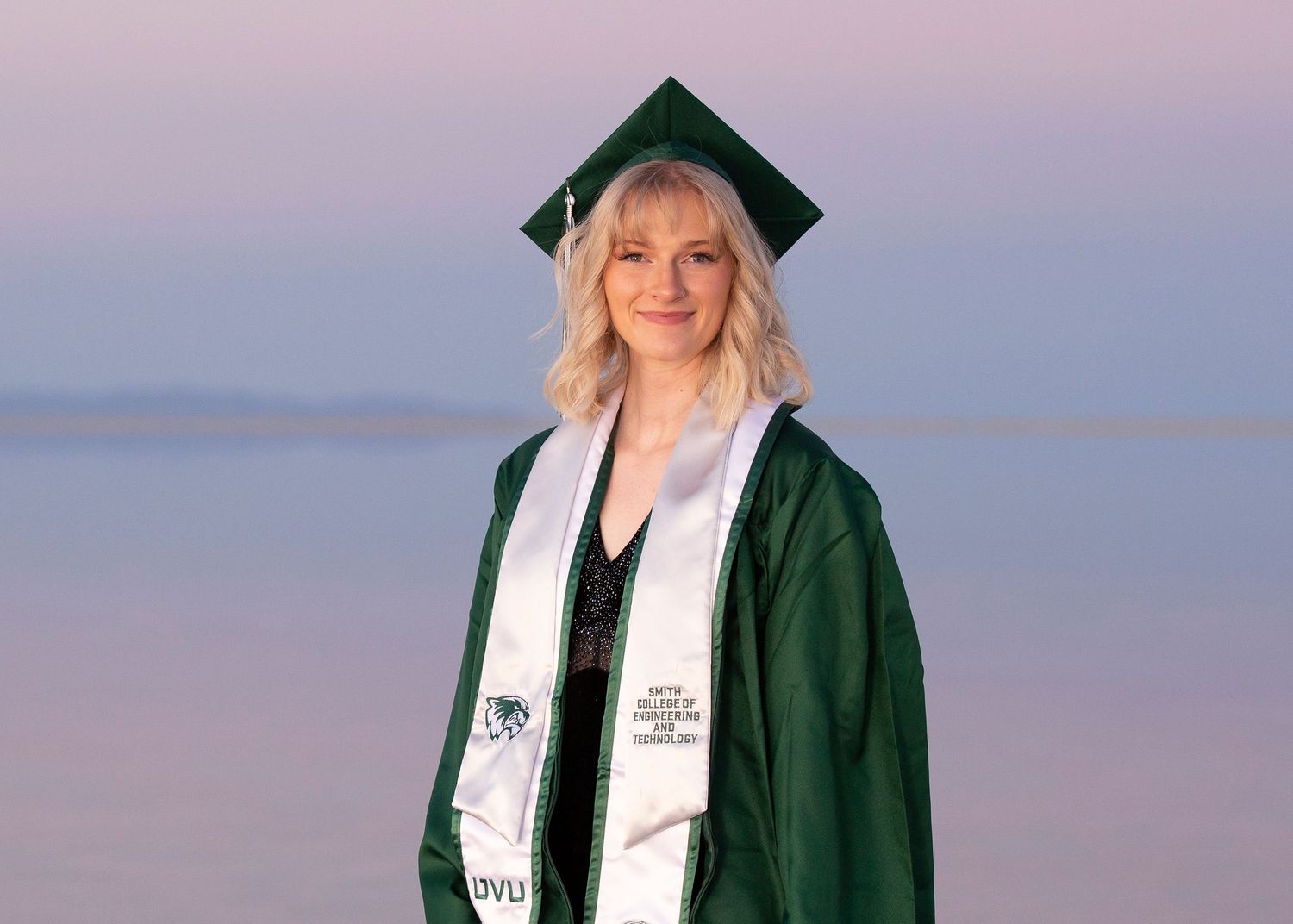 Graduate in green cap and gown with white graduation stole stands against soft sunset sky background. Bonneville Salt Flats Photography