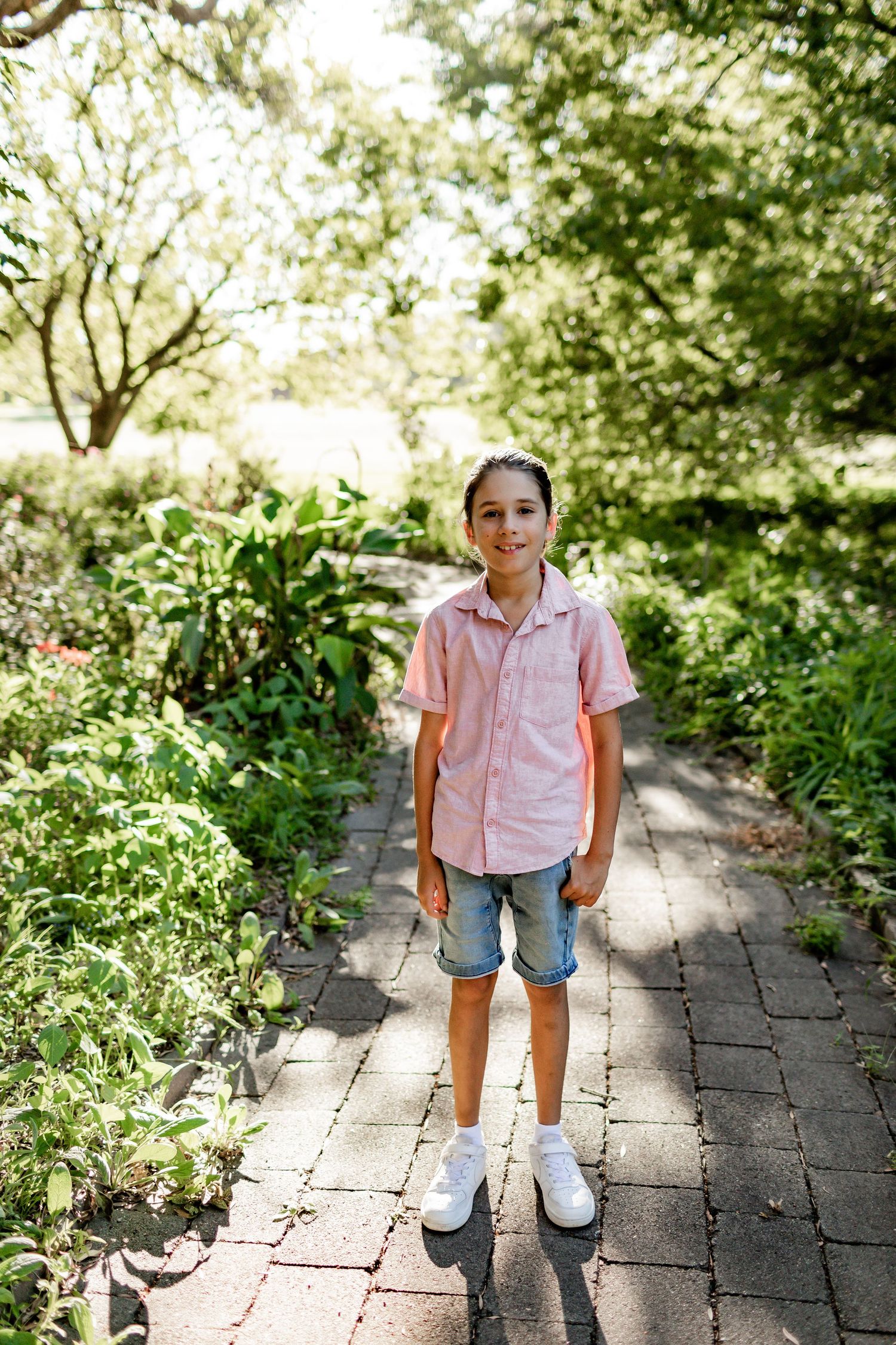 Person in pink shirt and denim shorts standing on brick pathway surrounded by greenery.