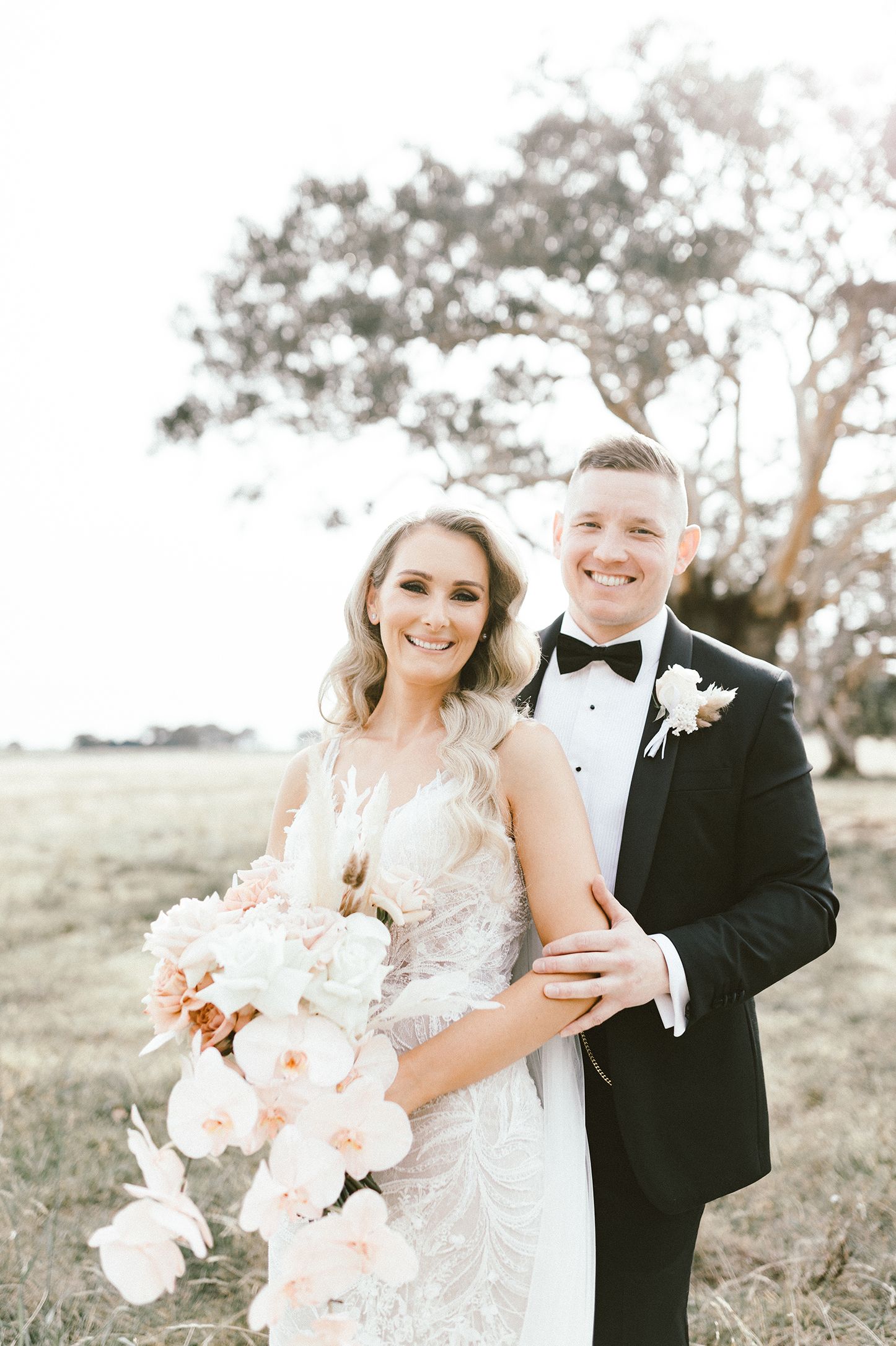 Wedding couple poses with peach and white floral bouquet against dreamy outdoor backdrop.