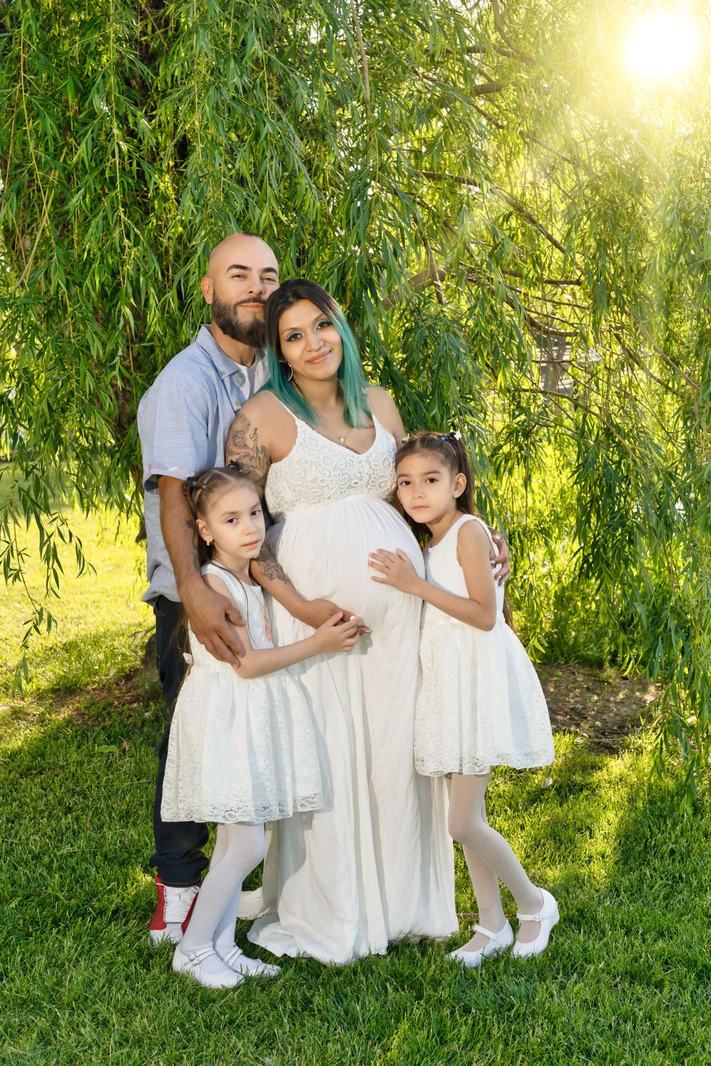A family poses under a willow tree in white outfits during a maternity photoshoot.