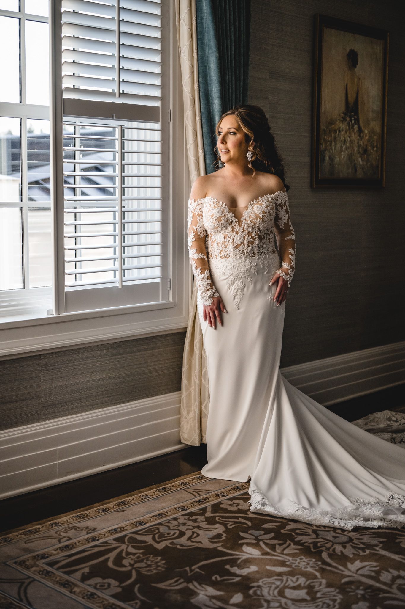 Elegant bride in off-shoulder lace wedding gown standing by window with plantation shutters.