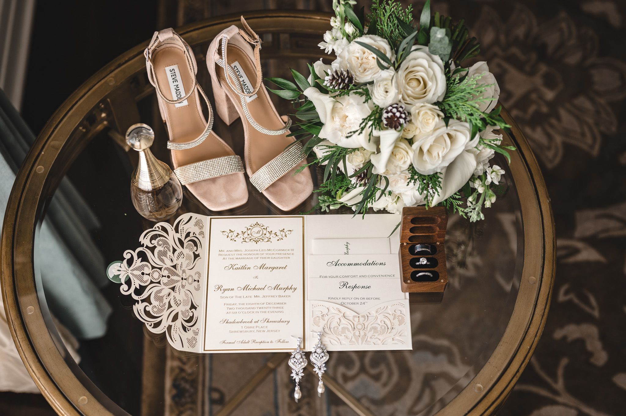 Wedding details flatlay with blush shoes, white floral bouquet and invitation on round table.