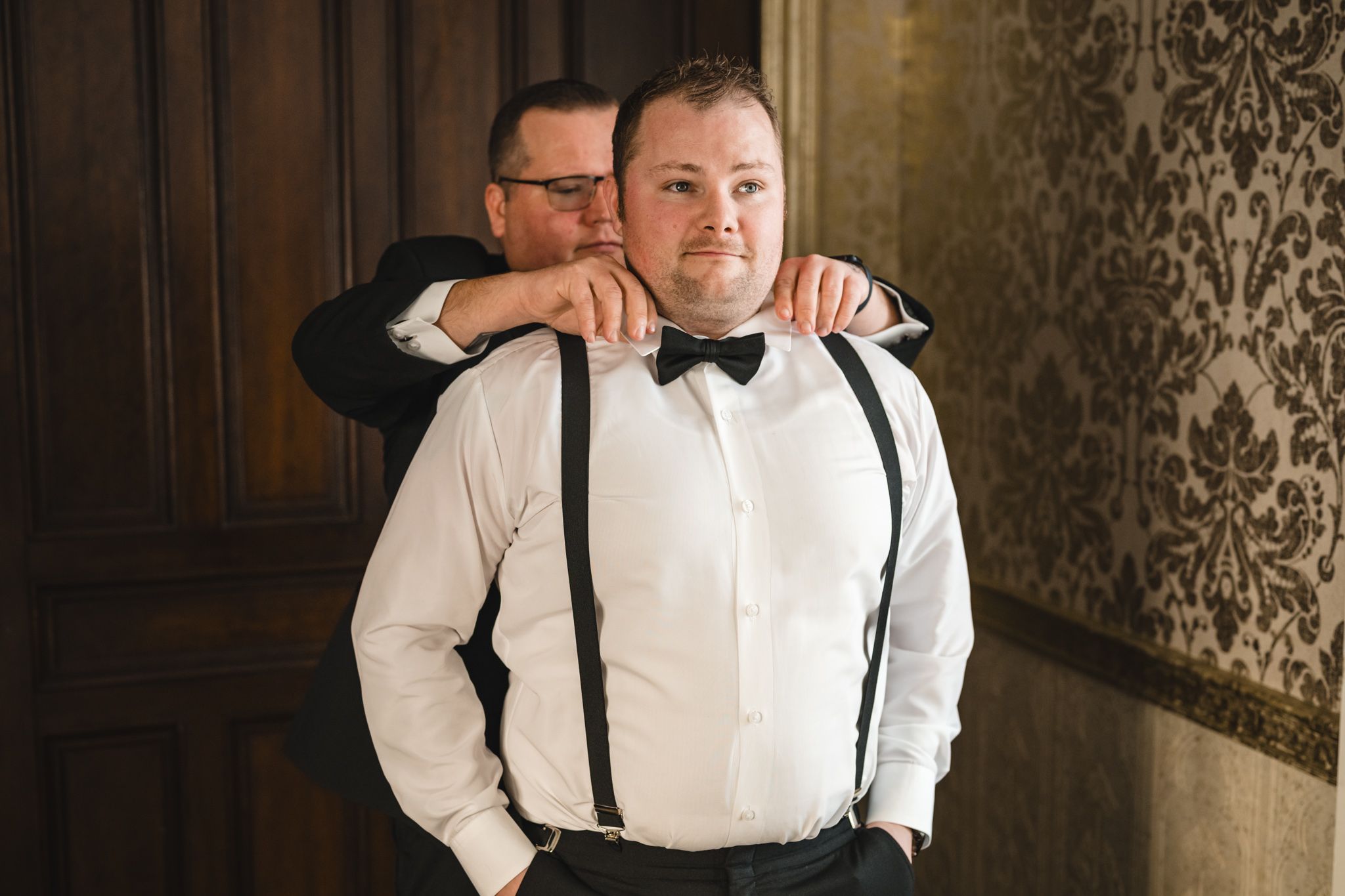 Two people share a lighthearted moment while adjusting formal wear near a window.