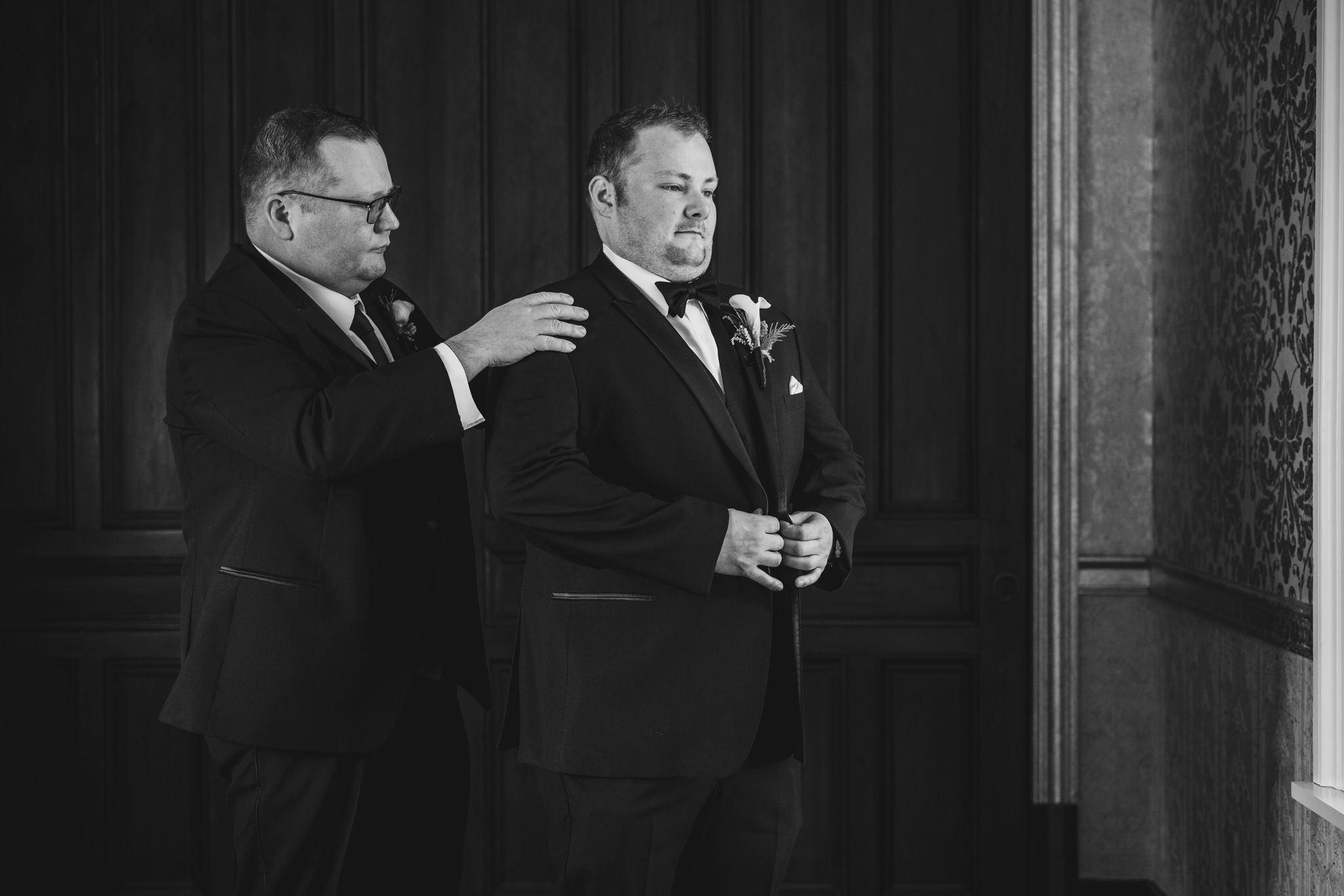 A black and white photo captures two people in tuxedos during wedding preparations.