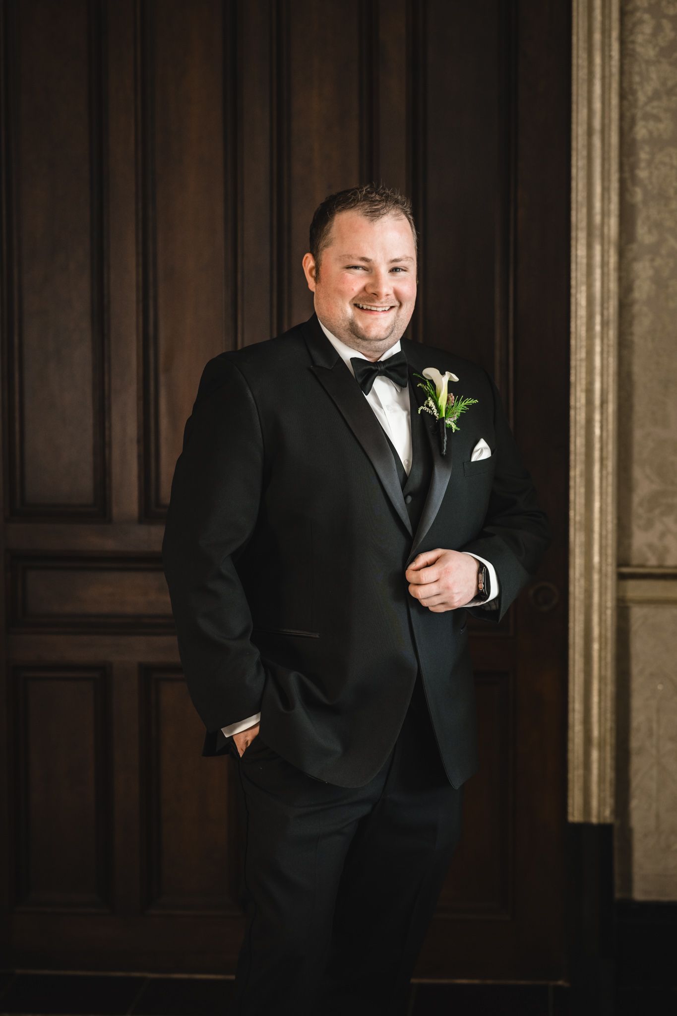 A person in a black tuxedo with a green boutonniere smiles in an elegant indoor setting.