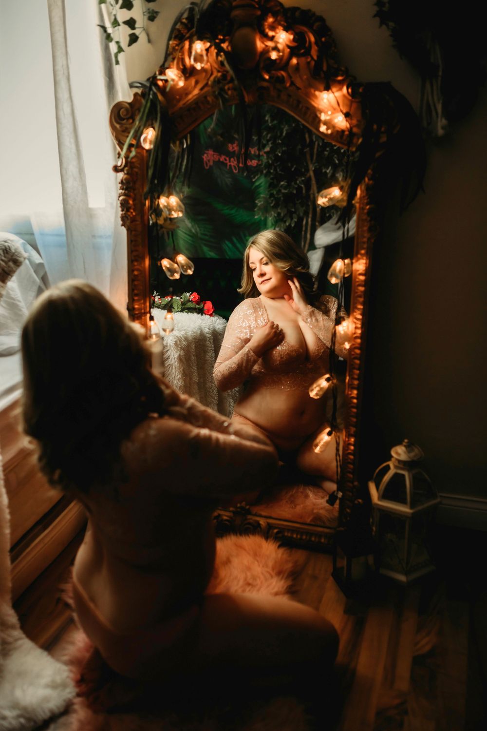 Mirror with ornate wood frame decorated with fairy lights reflects a soft and dreamy boudoir scene.