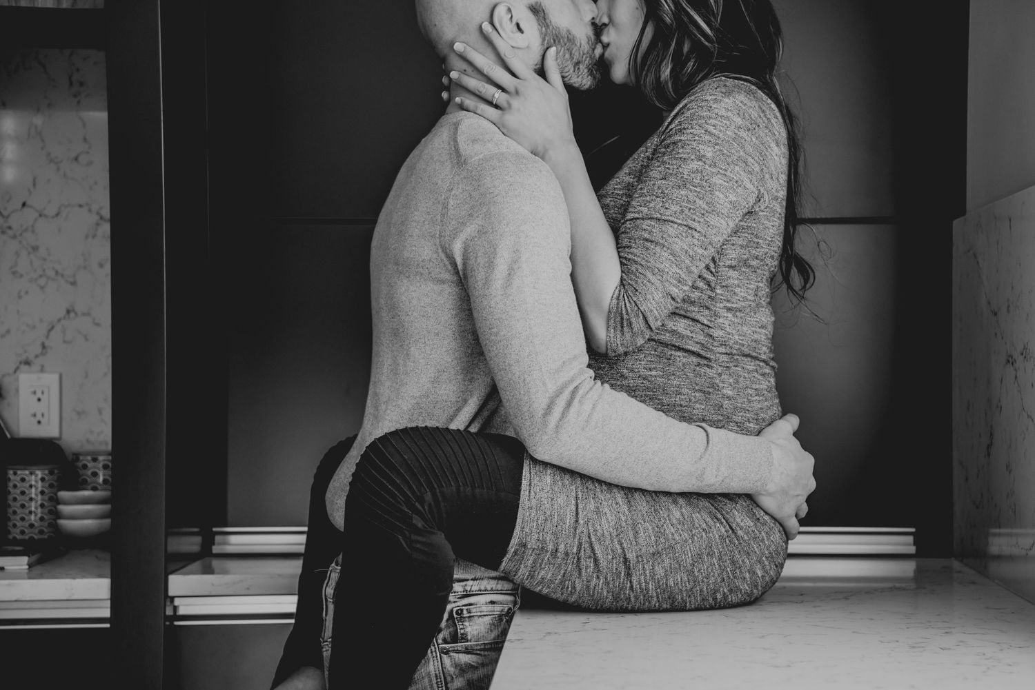 A tender black and white artistic photograph captures an intimate couple embracing while sitting indoors.