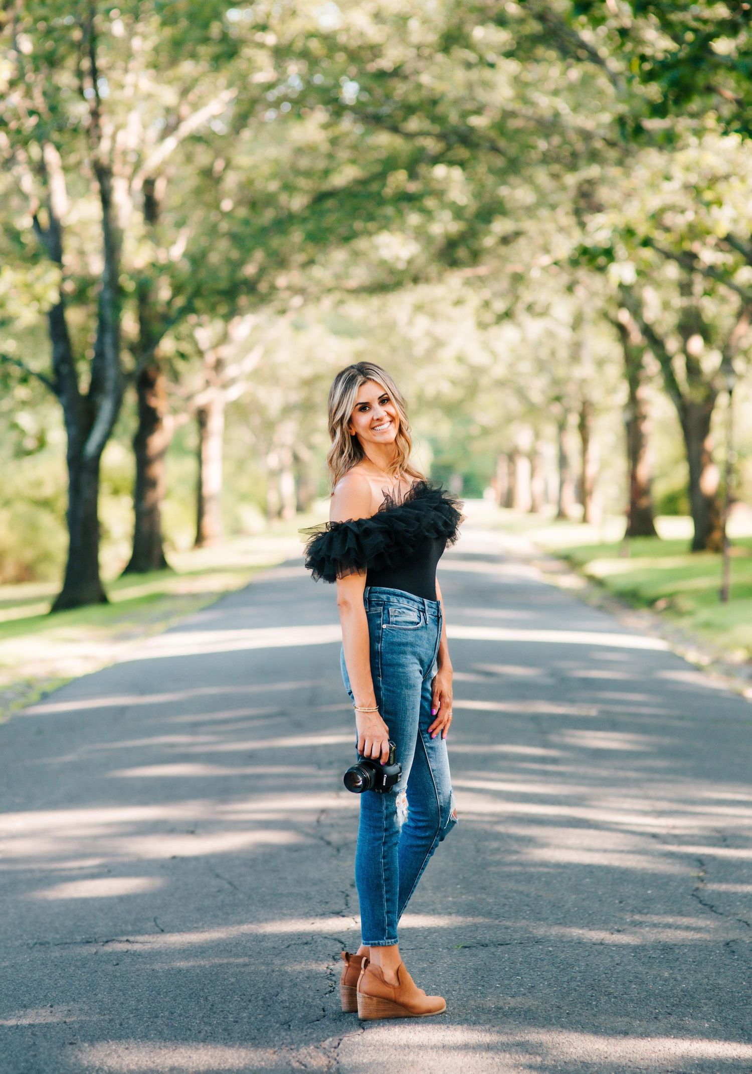 A woman in jeans and black ruffled top poses with camera on a tree-lined path.