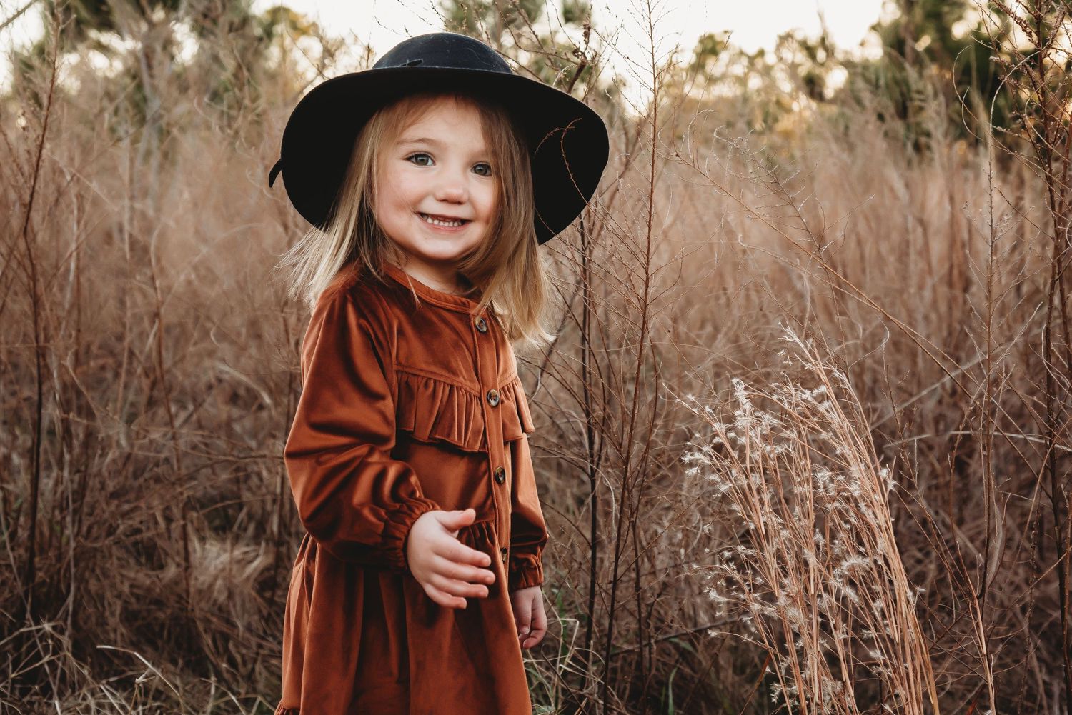 A child in a rustic brown dress and black hat stands joyfully among tall autumn grasses at golden hour.
