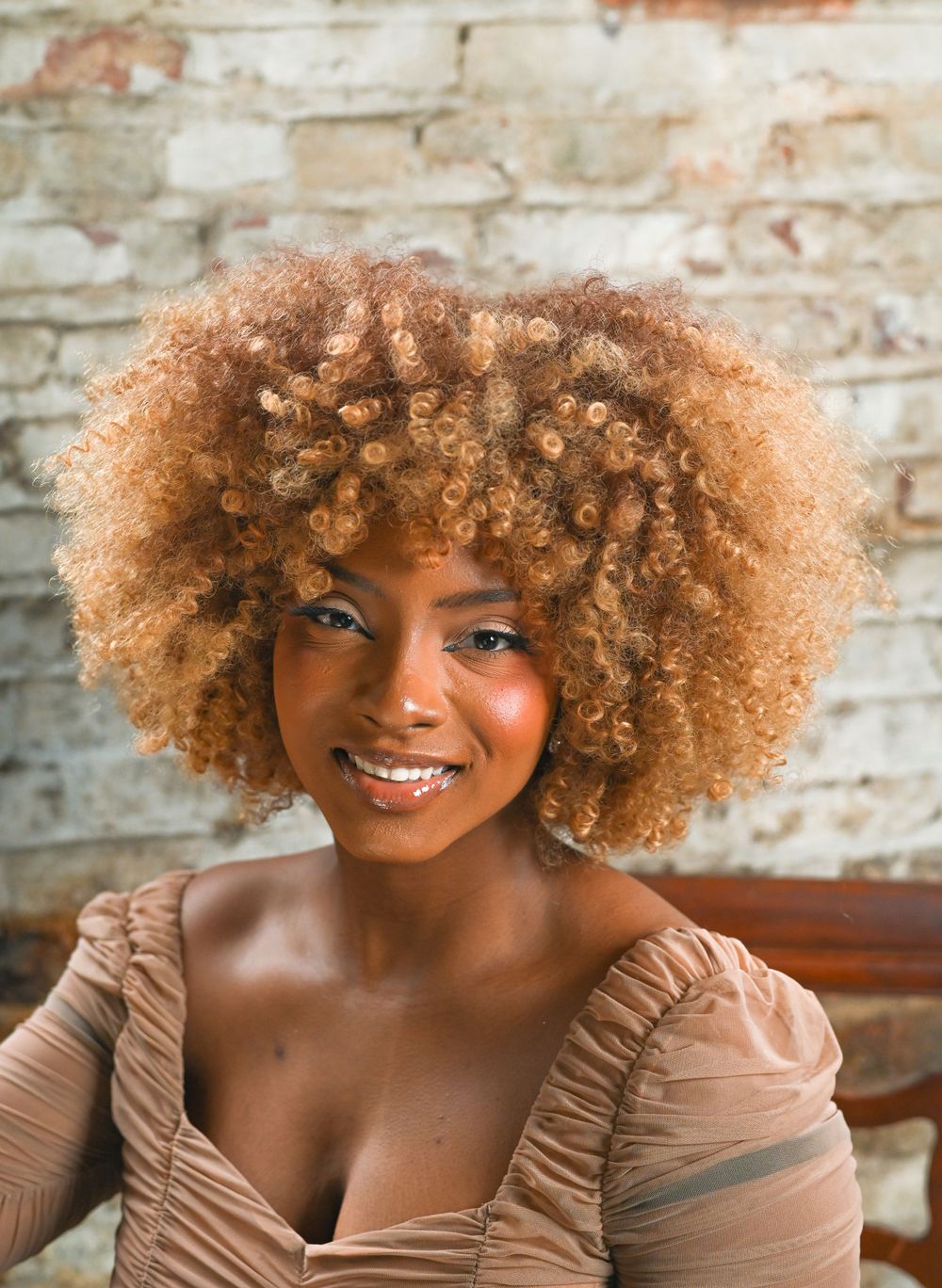 A glamour portrait with natural curly hair against a rustic brick wall background.