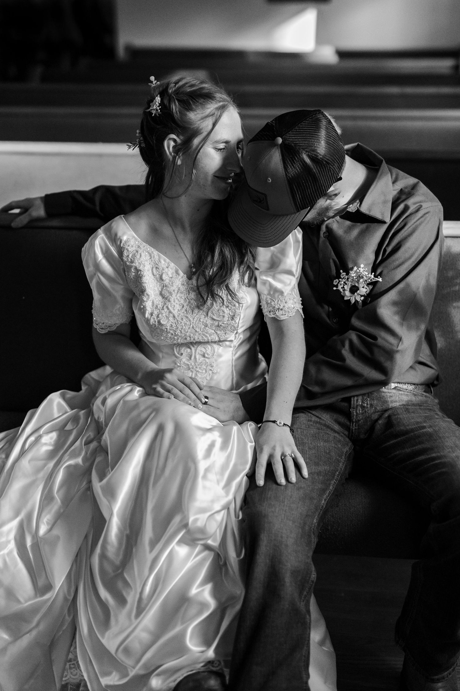 Black and white photo of a bride in white gown and groom in baseball hat sharing an intimate moment seated together.