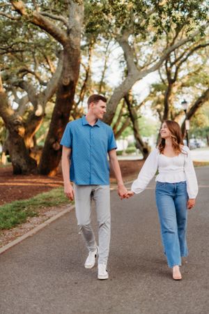 Couple holds hands while walking down tree-lined path in casual attire.