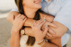 Close up of engaged couple embracing on beach showing rings and jewelry.