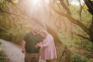 Couple embraces on a sunlit forest path surrounded by green trees and dappled light.