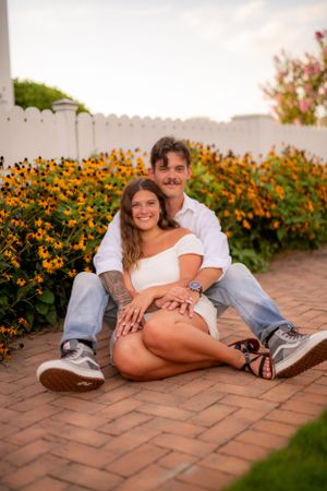 A casual couple relaxes on brick pavement with yellow flowers blooming behind them.