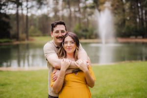 A romantic couple embraces by a lake with a fountain in the background.