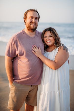 Couple posing on beach at sunset wearing casual attire with ocean in background.