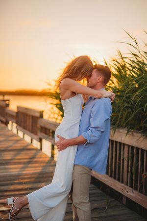 Romantic sunset photo of couple embracing on wooden boardwalk by water.