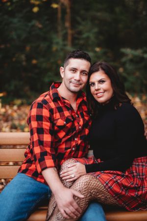 A couple in matching buffalo plaid and black outfits sits together on a wooden bench during autumn.