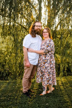 Couple poses together under willow tree in coordinated casual outfits.