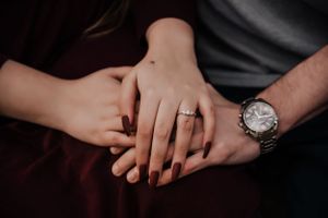 Close up of hands with engagement ring and wristwatch on dark fabric.