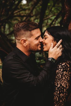 Romantic close-up moment between couple in formal black attire against forest backdrop.