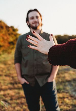 A hand showing off an engagement ring with a blurred figure in the background.