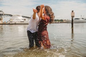 Two people splashing and playing in water by marina with boats in background.