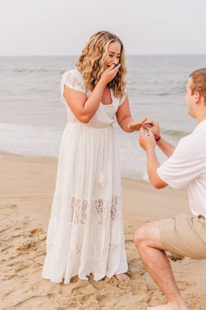 Woman in white lace dress reacts emotionally to beach proposal.