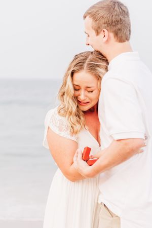 Romantic beach proposal moment with white dress and ring box at sunset.