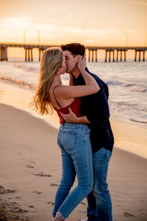 Couple embracing and kissing on sandy beach at sunset with pier in background.