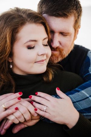 Engaged couple in casual winter wear embrace during romantic photoshoot with visible diamond ring.