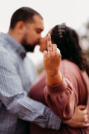 Woman showing off engagement ring while couple shares intimate moment in background.