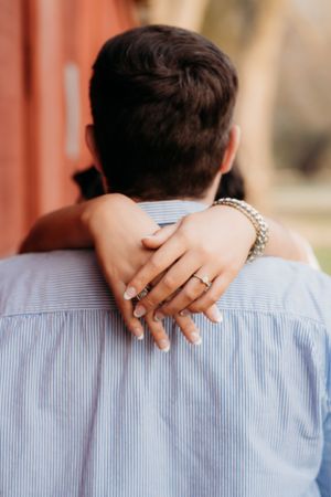 Close-up of hands embracing from behind, showing an engagement ring against a blue striped shirt.