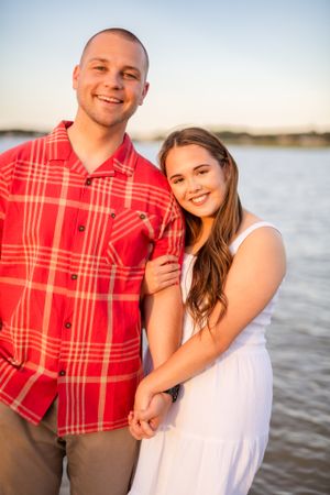 Waterfront portrait of couple in coordinated outfits of white dress and red plaid shirt.